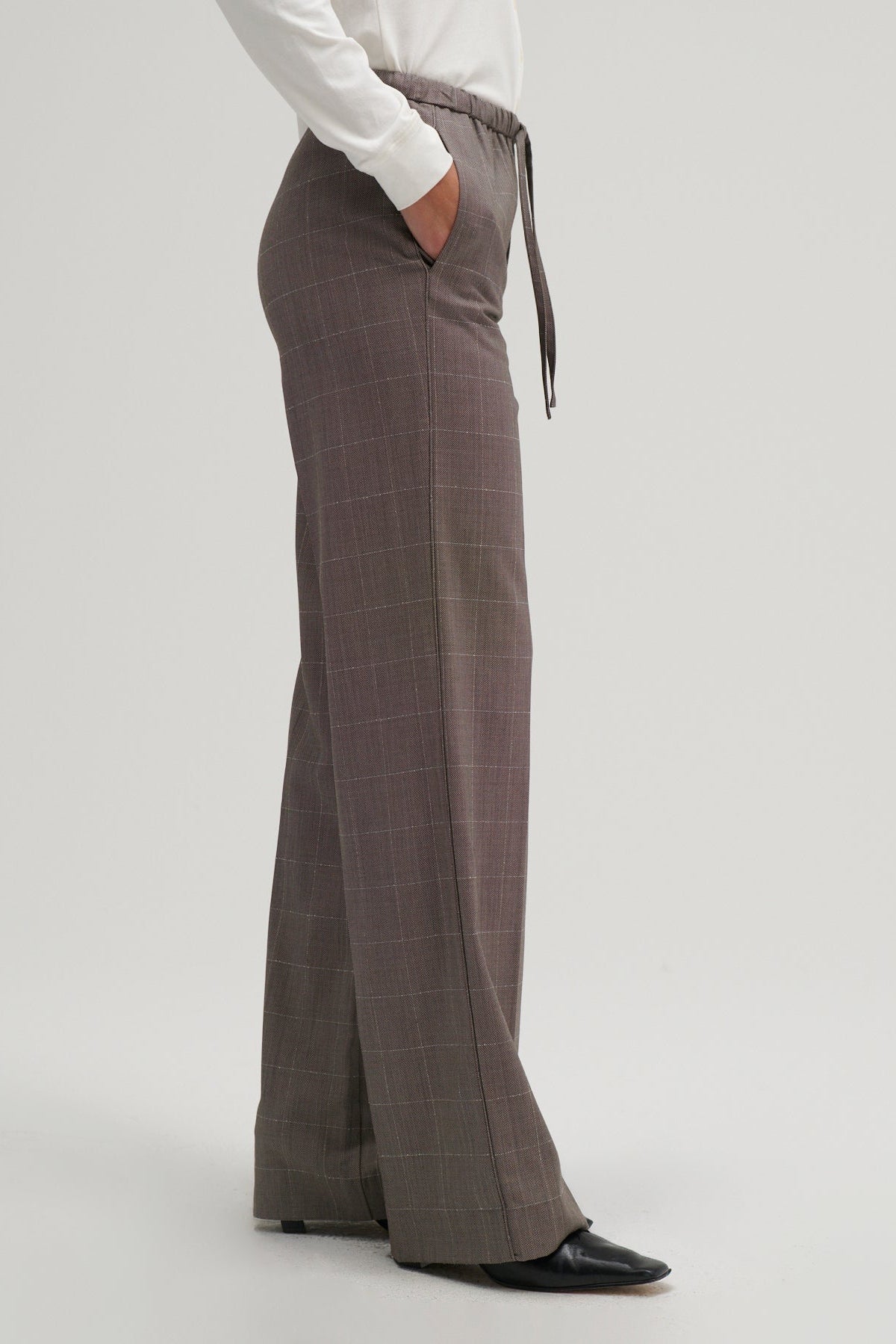 A person wearing high-waisted, wide-leg trousers in a muted brownish-gray plaid pattern, styled with a white long-sleeve top and black shoes against a light gray studio background.