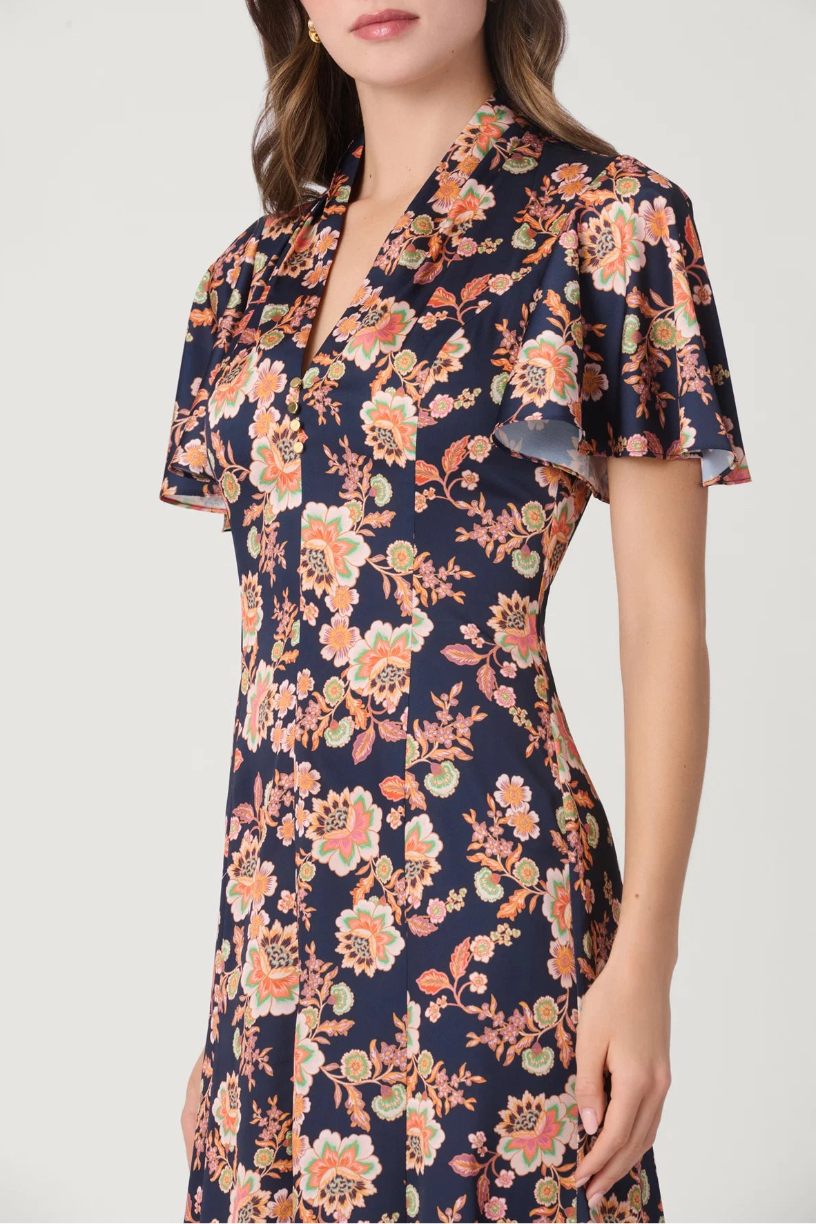 A woman wearing a navy blue short-sleeve dress with a vibrant orange, peach, and cream floral print. The dress features a split V-neckline with ruffled trim and a fitted silhouette.