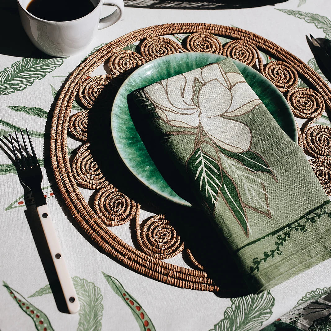 A styled table setting featuring a sage green linen napkin with a large white magnolia floral print, layered on a green ceramic plate and a brown woven placemat. A white mug, a cutlery handle, and a matching botanical print tablecloth are visible.