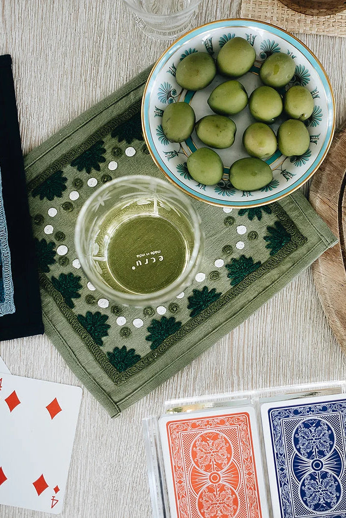 An overhead styled flat-lay featuring a bowl of green olives, a glass of white wine, playing cards (including Bicycle cards), nuts, and cured meats on a light wood background.