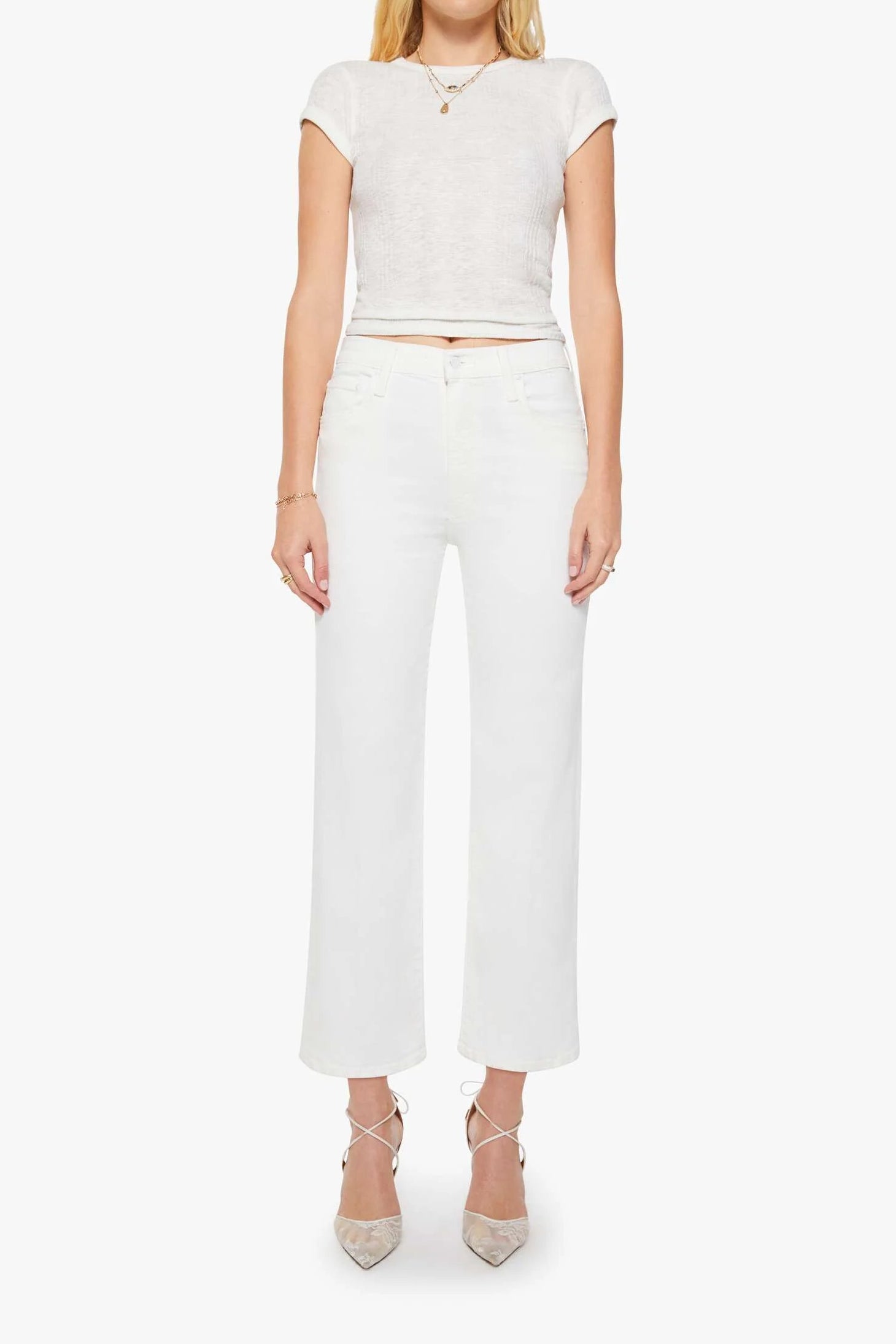 A blonde model stands against a light gray studio background wearing a white short-sleeve cropped knit top, white wide-leg cropped pants, and beige pointed-toe heels.