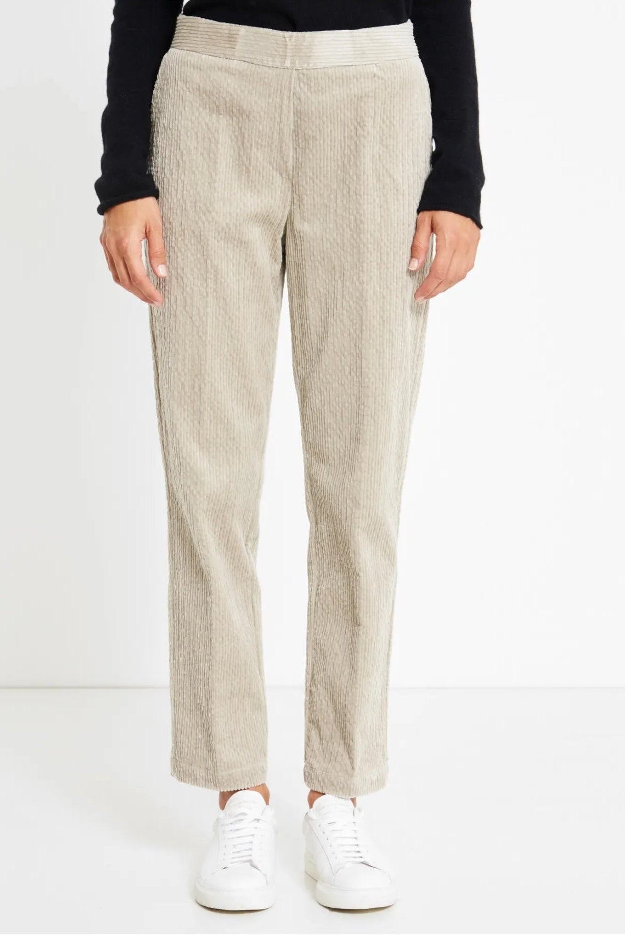 A person wearing textured light beige straight-leg trousers, a black long-sleeve top, and white sneakers, standing against a clean white studio background.