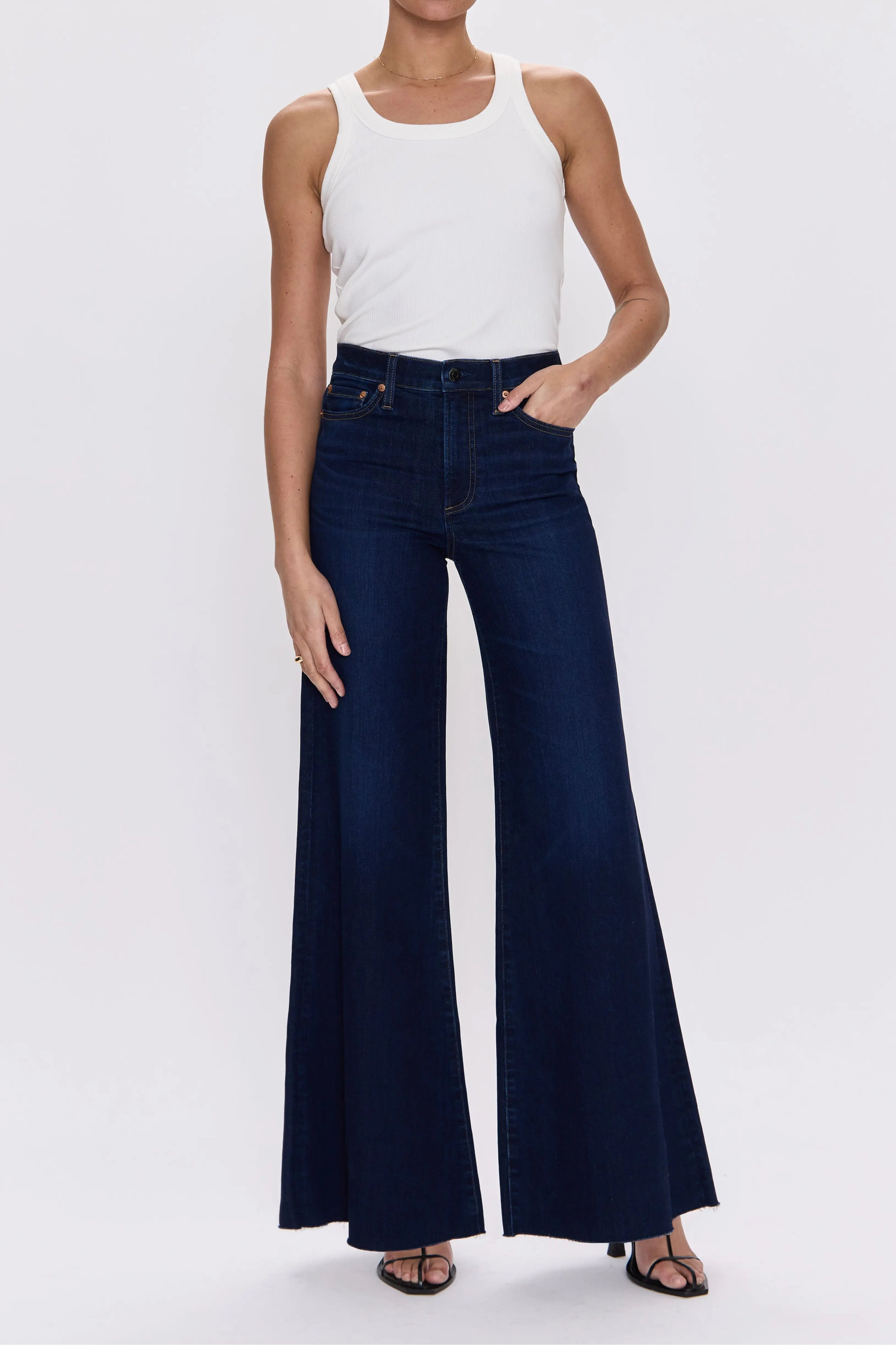 A model wearing dark blue wide-leg flare jeans with a white tank top, standing against a light gray studio background.