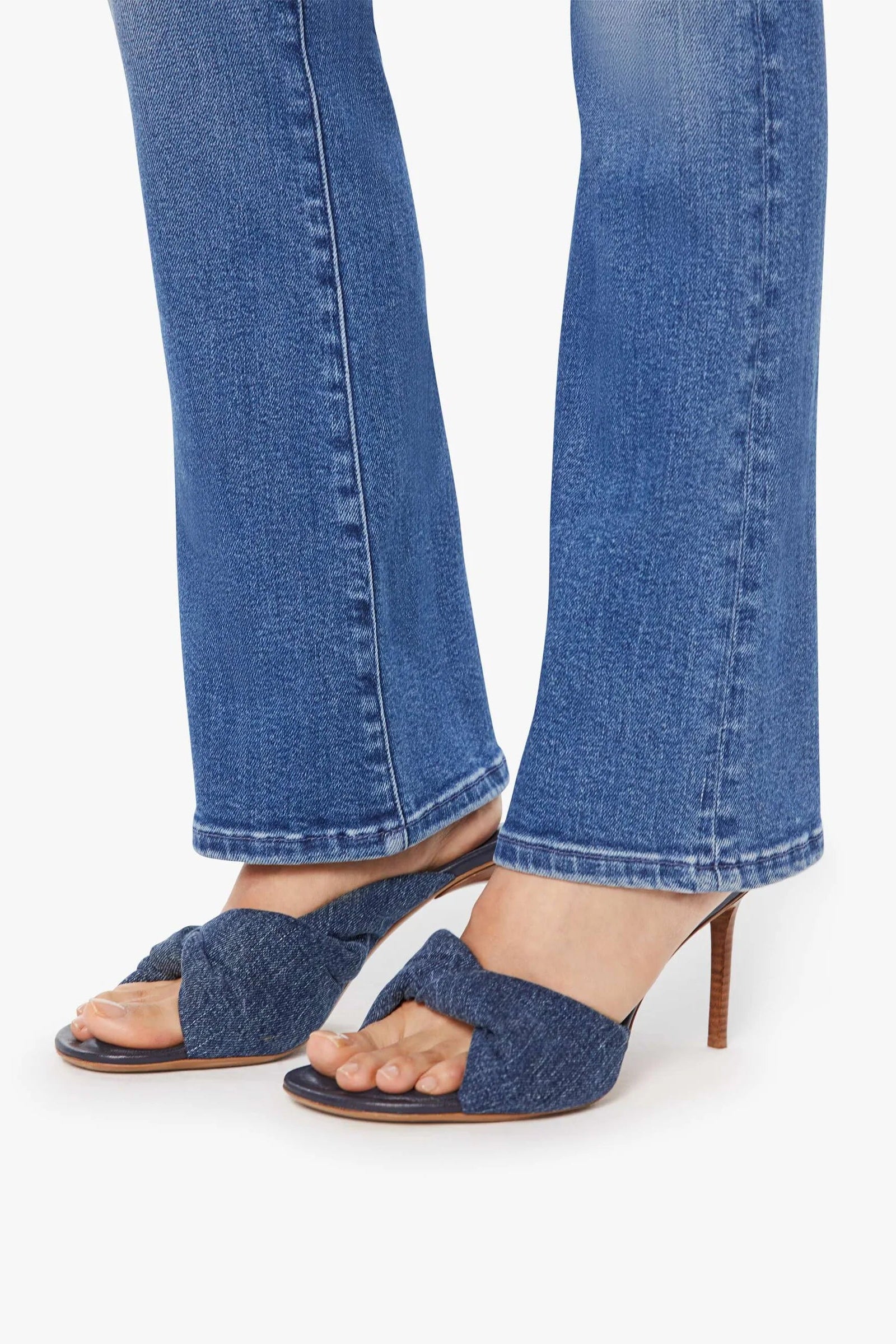 A close-up of a person's feet wearing blue denim heeled sandals with wide crisscross straps and blue denim flare jeans, set against a plain white background.