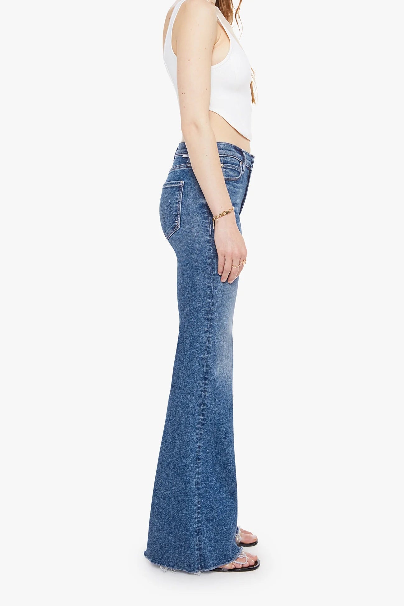 A side-profile view of a model wearing blue high-waisted flare jeans, a white crop top, and sandals against a plain white background.