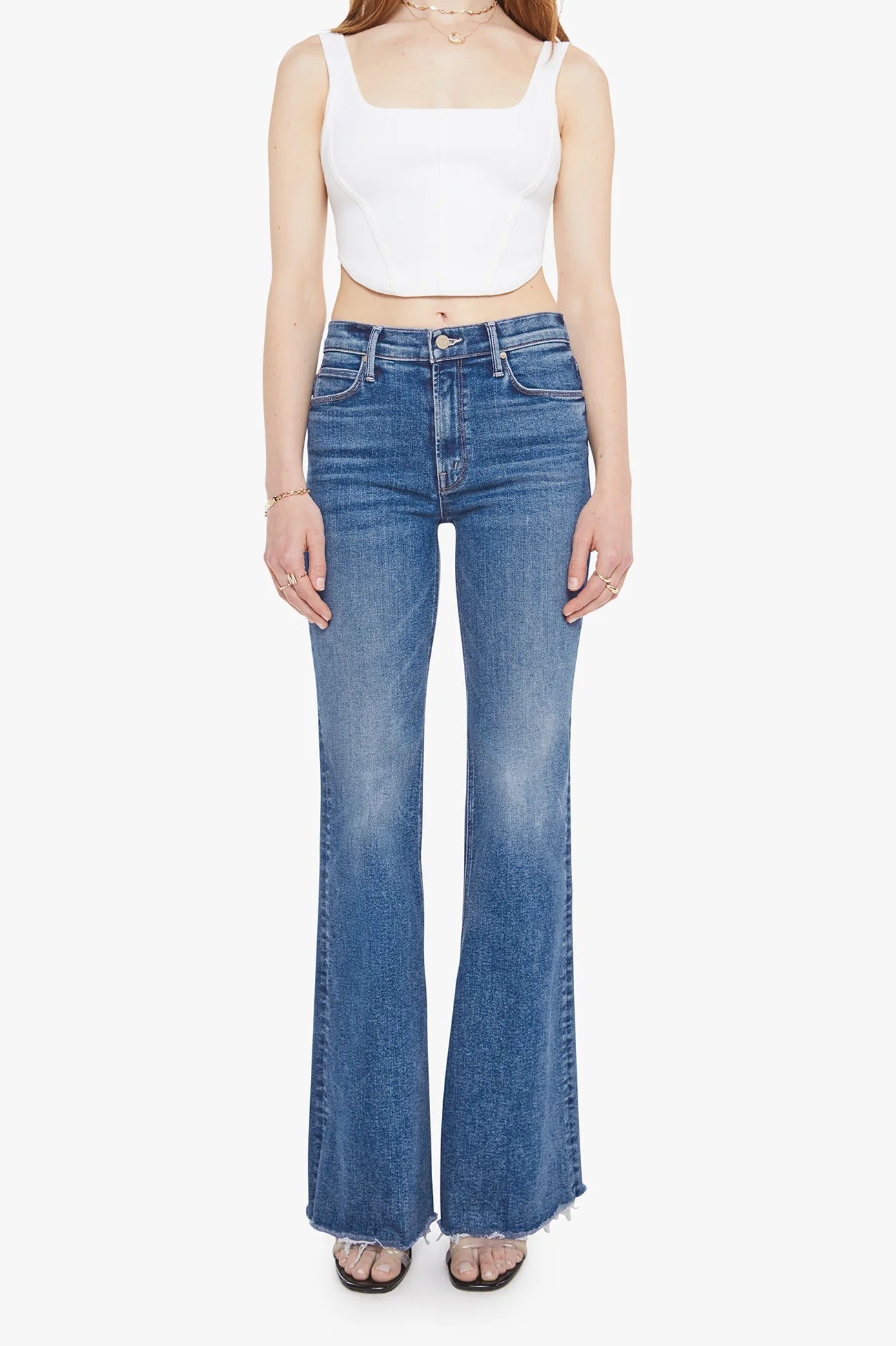A model wearing a pair of medium-wash blue flare jeans with a frayed hem, paired with a white crop top and flat sandals, against a plain white background.