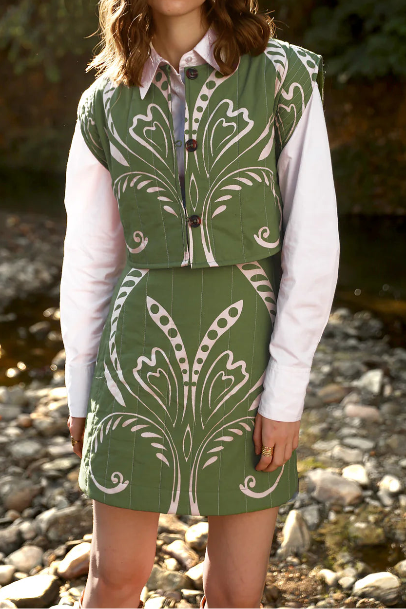 Woman wearing a green patterned vest and skirt standing outdoors near a stream.