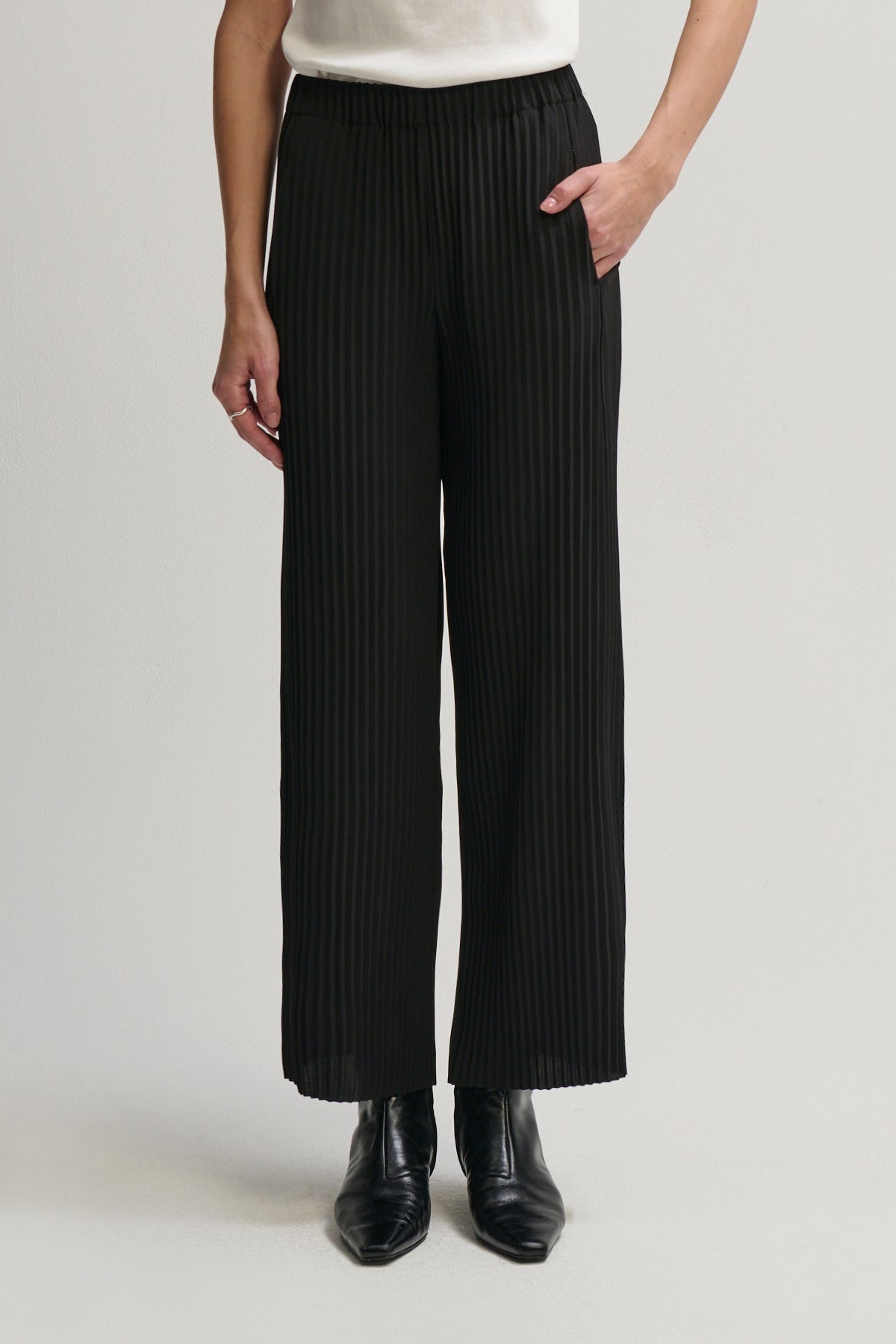 A model wearing black pleated wide-leg trousers with an elastic waistband, paired with a white top and black leather ankle boots, shown against a light gray studio background.