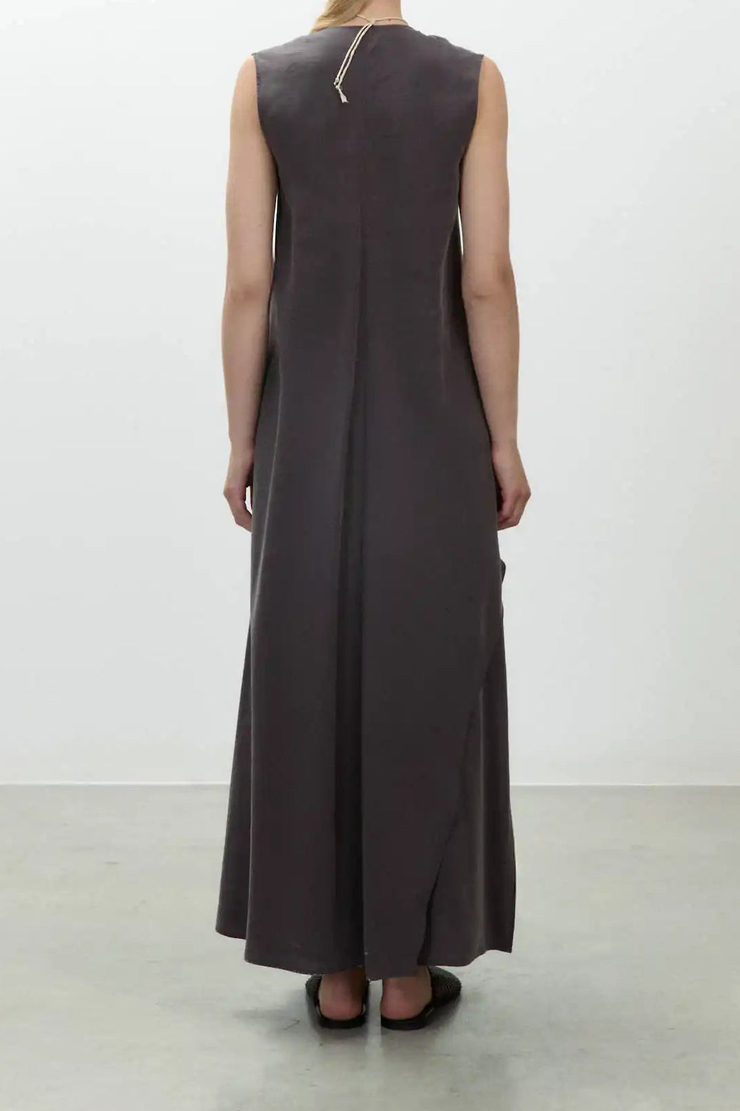 A woman stands with her back to the camera wearing a sleeveless, floor-length dark charcoal dress in a minimalist studio with a white wall and gray floor.