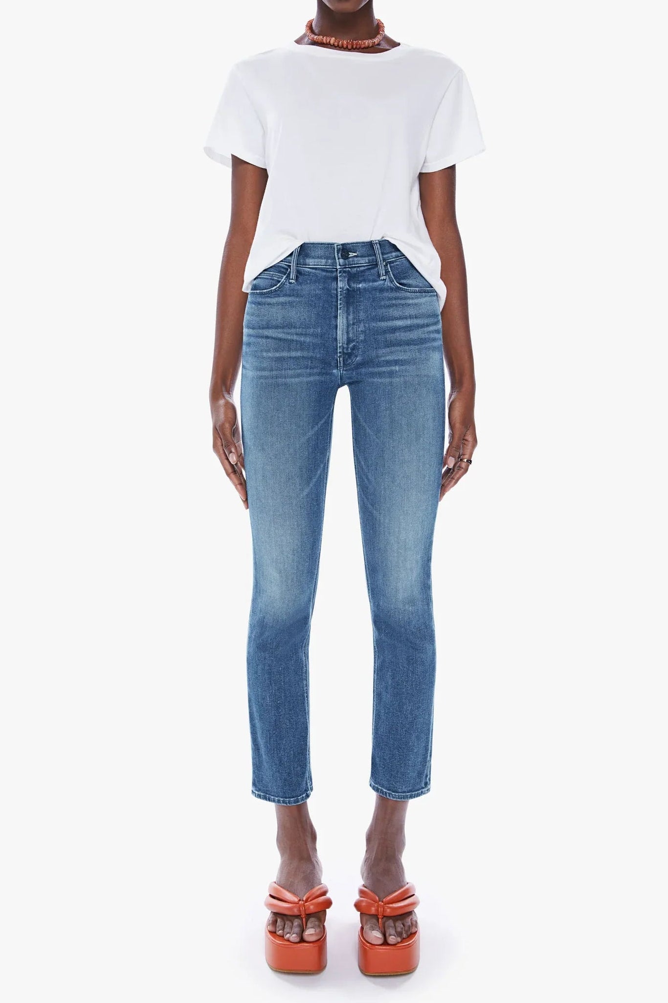 A model stands against a light gray studio background wearing a white short-sleeve top, blue cropped jeans, and orange platform sandals.
