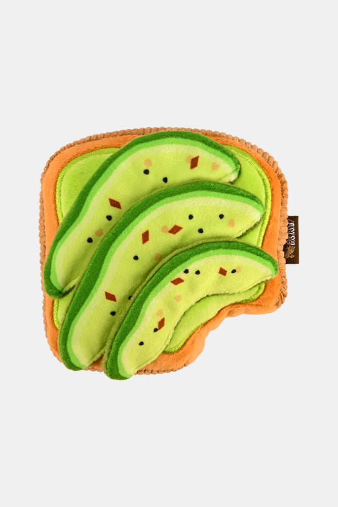 A plush avocado-shaped cushion or pillow on a plain white background. The design features light green and dark green avocado slices with small embroidered black seeds and red-orange seed details, set on an orange-brown rind-like border.