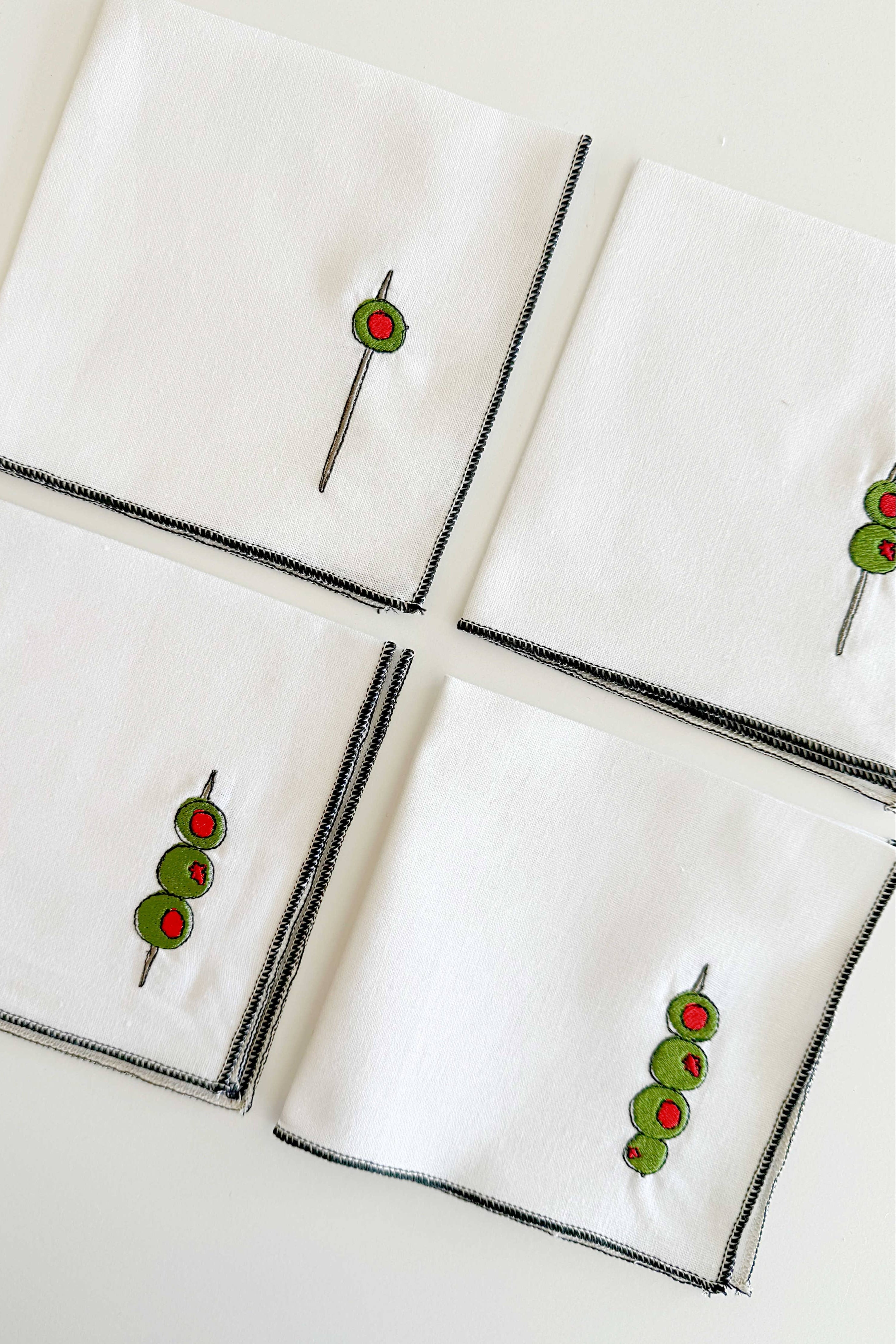 Set of white napkins with black borders featuring embroidered green olives on a white background
