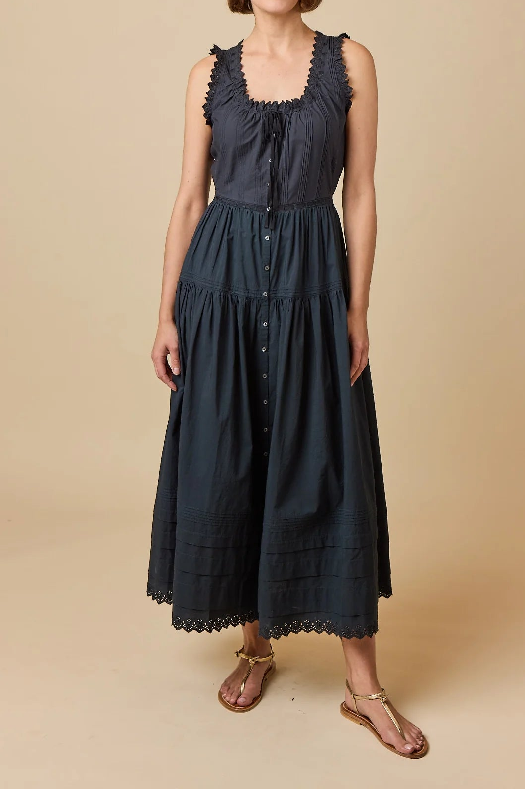A woman modeling a sleeveless dark navy midi dress with a gathered bodice, button front, and scalloped trim, standing against a solid beige studio background.
