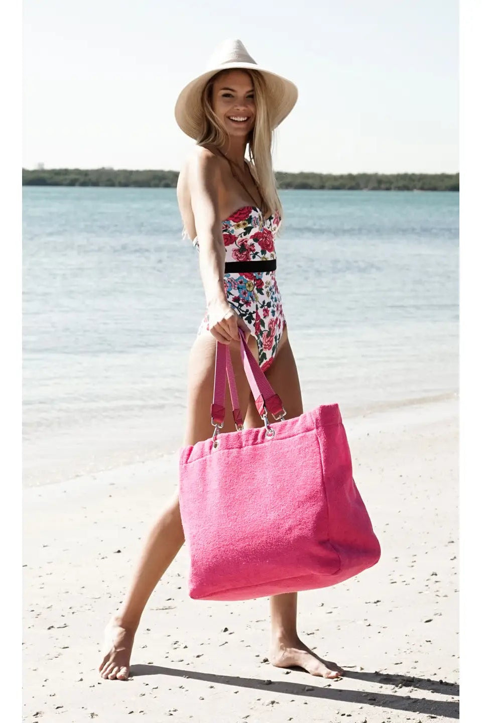 Woman on a beach holding a pink tote bag, wearing a floral swimsuit and sun hat.