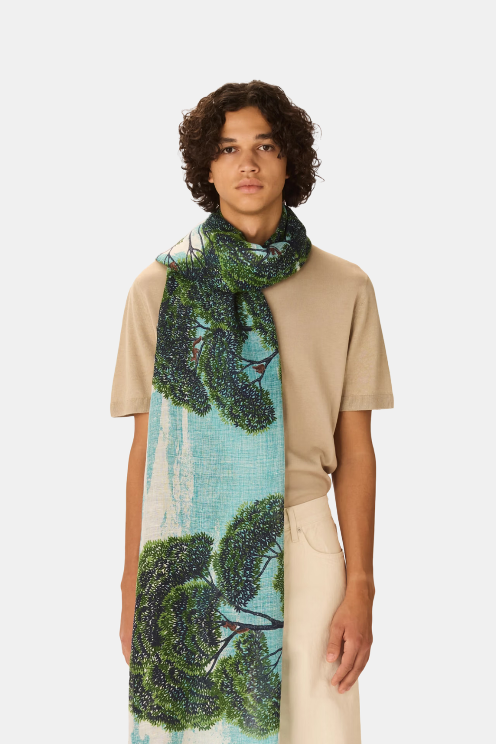 A model wearing a beige short-sleeve top and cream trousers, draped with a long scarf featuring a green tree pattern on a light blue background, against a light gray studio backdrop.