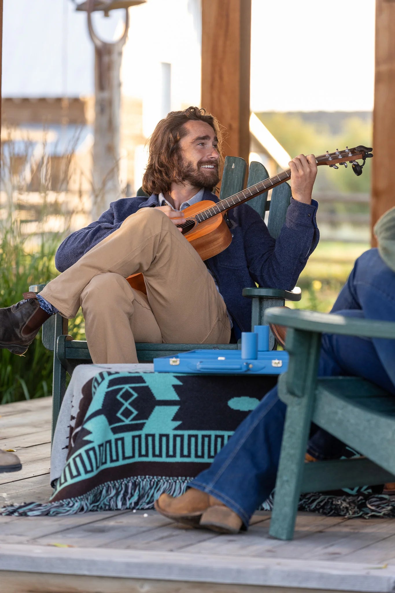 Man playing guitar on a wooden deck with a blue blanket and green chair.