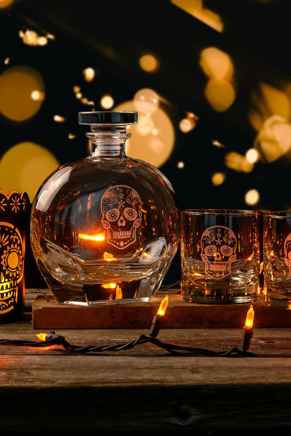 Glass decanter and skull-themed glasses on a wooden surface with blurred lights in the background