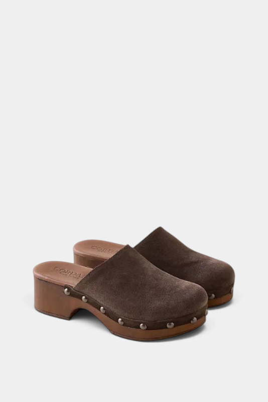 A pair of dark brown suede clogs with wooden block heels and silver metal studs, shown on a light beige background.