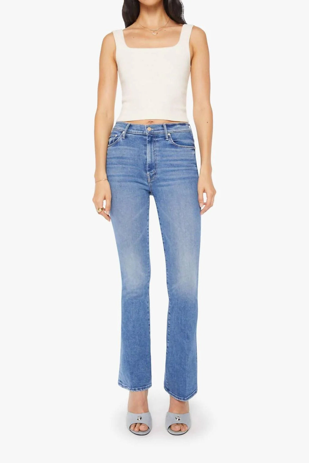 A model wearing light-wash blue flare jeans, a white ribbed tank top, and gray heeled sandals, standing against a plain white studio background.