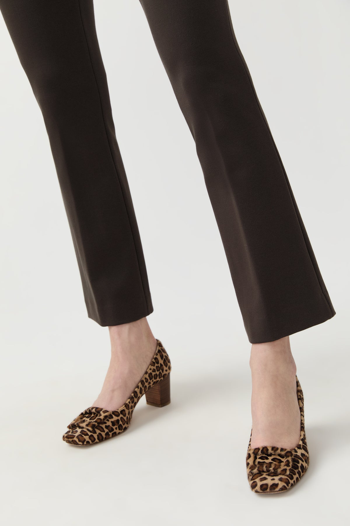 A person wearing dark brown flared trousers and leopard-print block-heel pumps, shown from the calves down against a clean light gray studio background.