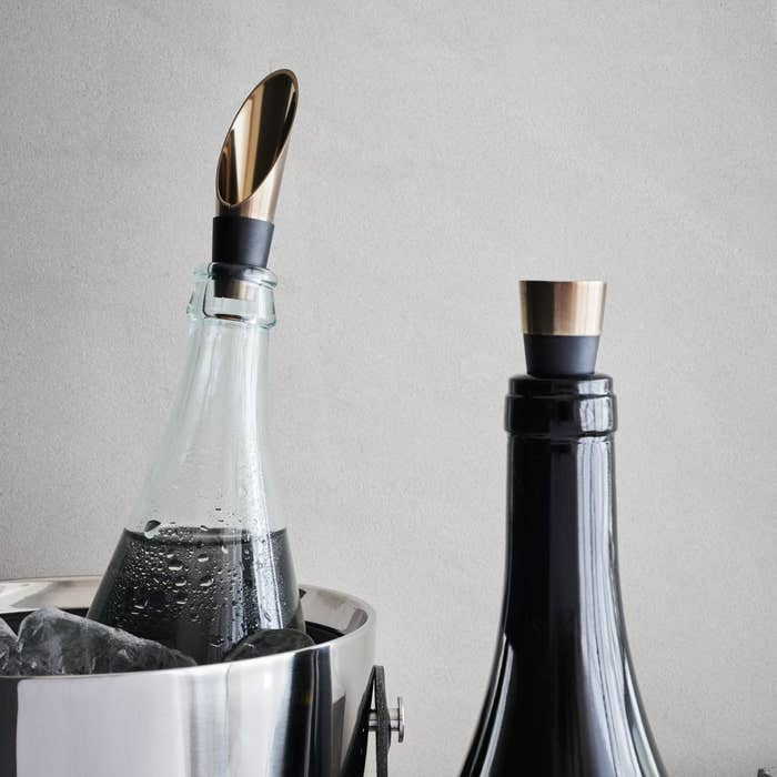 Two wine bottles with gold bottle stoppers, one in an ice bucket, on a neutral background.