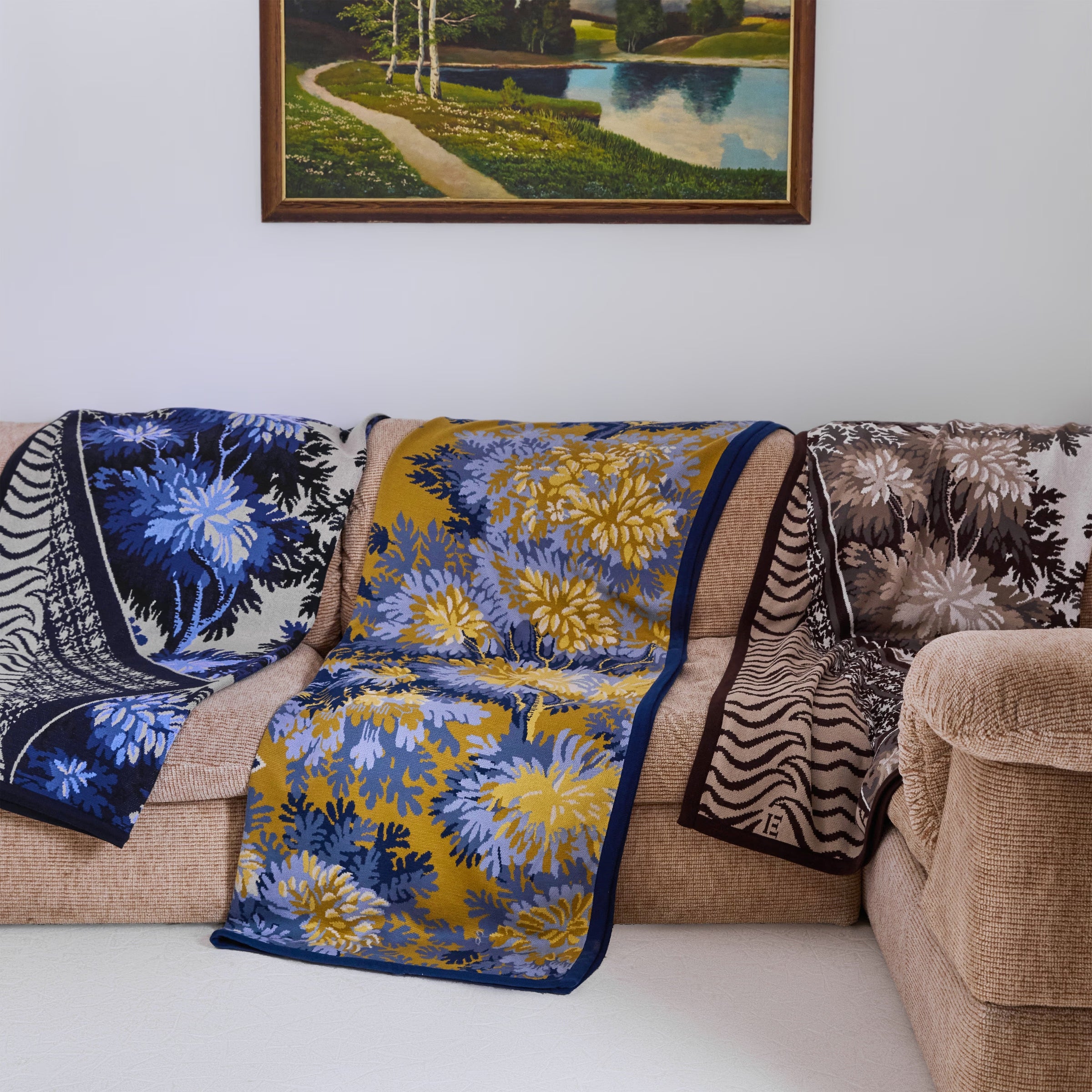 Beige sofa with floral-patterned blue and yellow blanket, zebra print blanket, and landscape painting on wall.