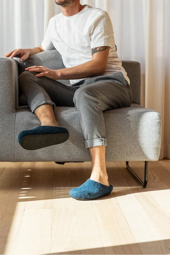 A man sits in a light gray upholstered armchair in a bright room with sheer curtains and wood flooring, wearing a white T-shirt, gray pants, and blue slip-on shoes.