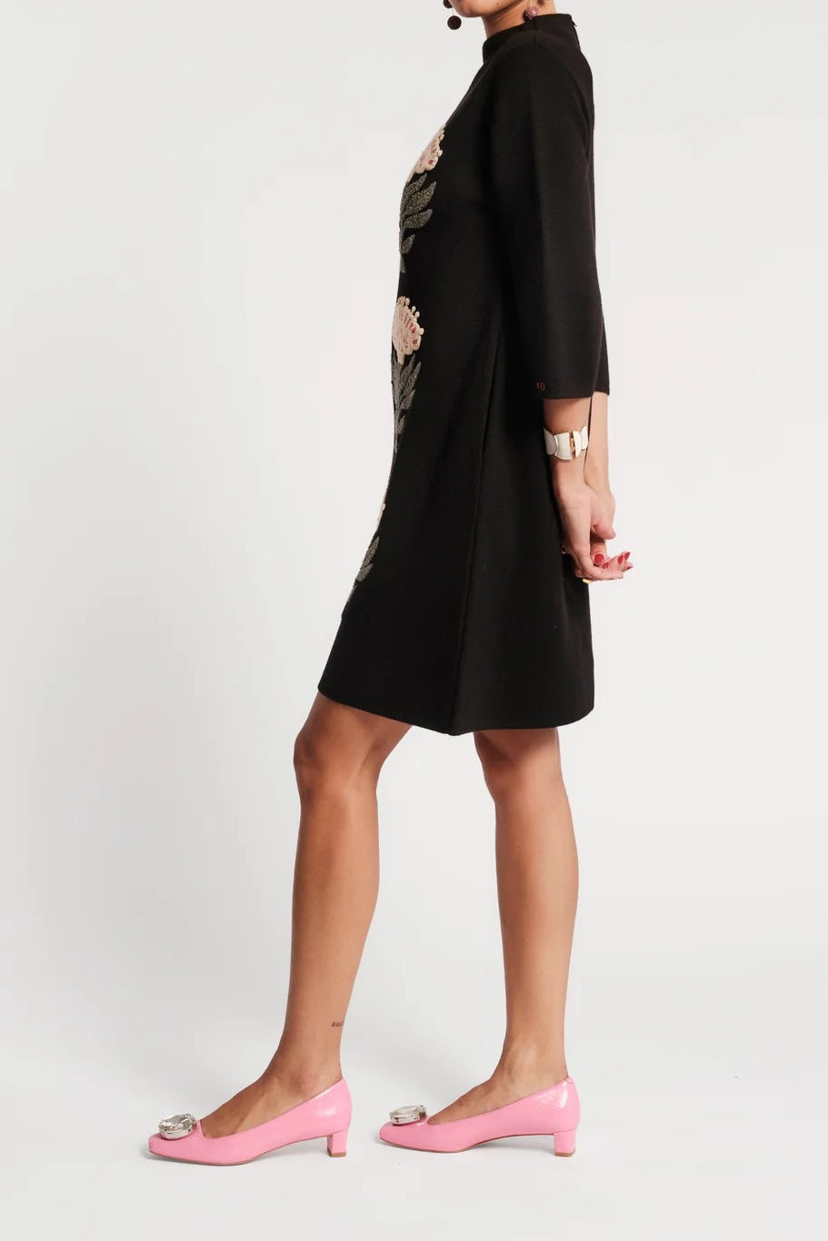 A woman modeling a black knee-length dress with three-quarter sleeves and side cutouts, paired with pink kitten heels, photographed against a plain light gray studio background.