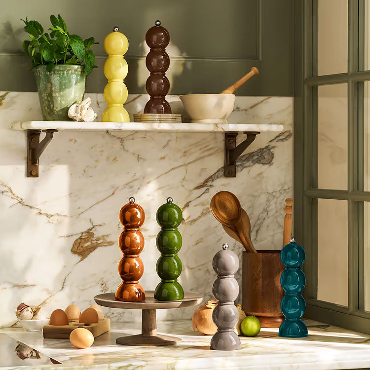 A styled kitchen scene with multiple decorative stacked ceramic pepper mills in assorted colors including yellow, brown, green, orange, gray, blue, and teal, arranged on a marble countertop and shelf.