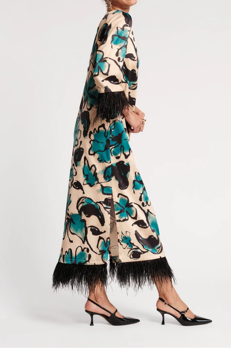 A woman modeling a long beige maxi dress with a bold teal, black, and white floral pattern and black feather fringe at the hem and cuffs, paired with black slingback heels against a light gray studio background.