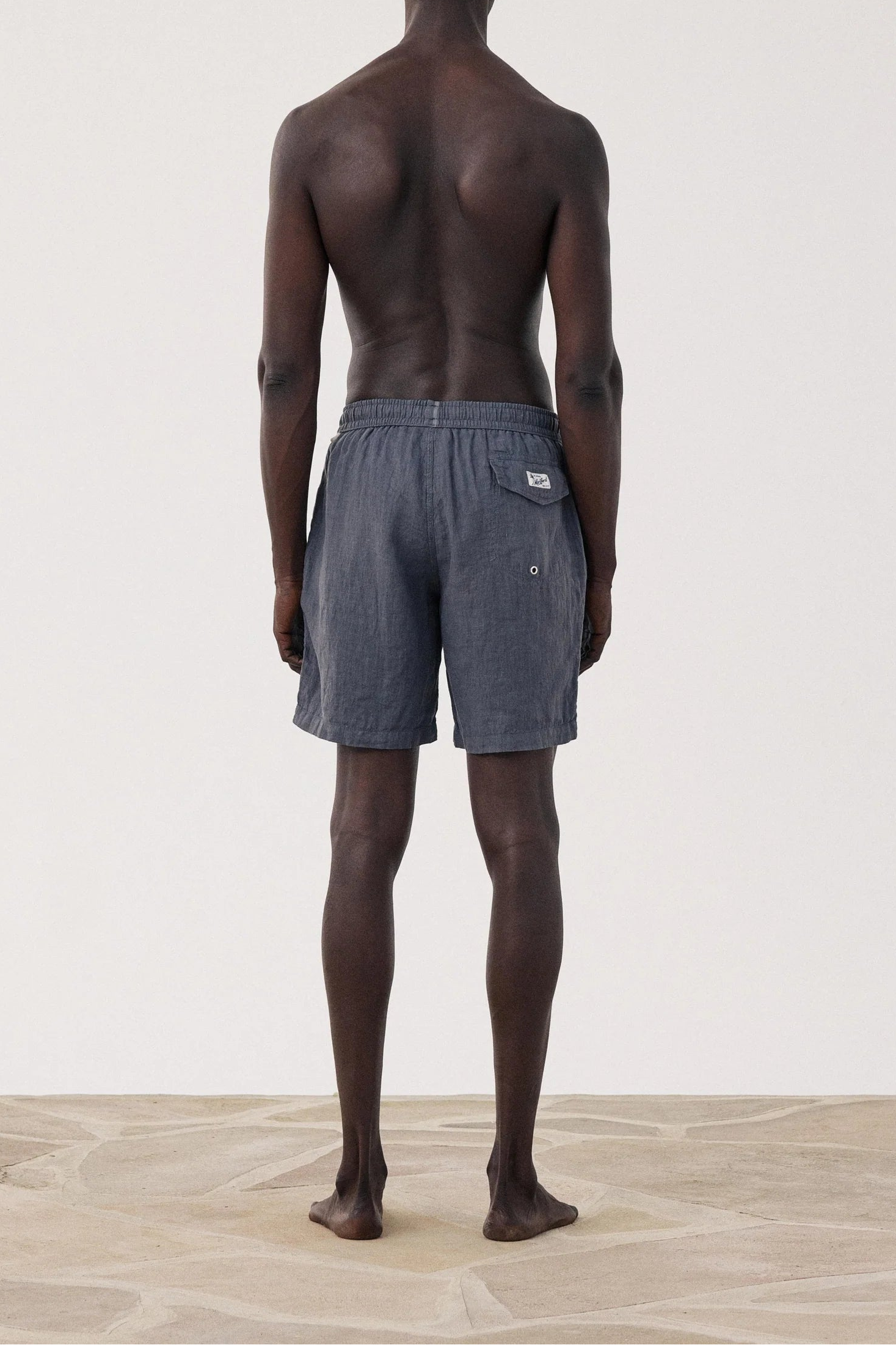 A shirtless man stands with his back to the camera wearing dark gray drawstring shorts, photographed in a minimalist studio with a light beige floor and wall.