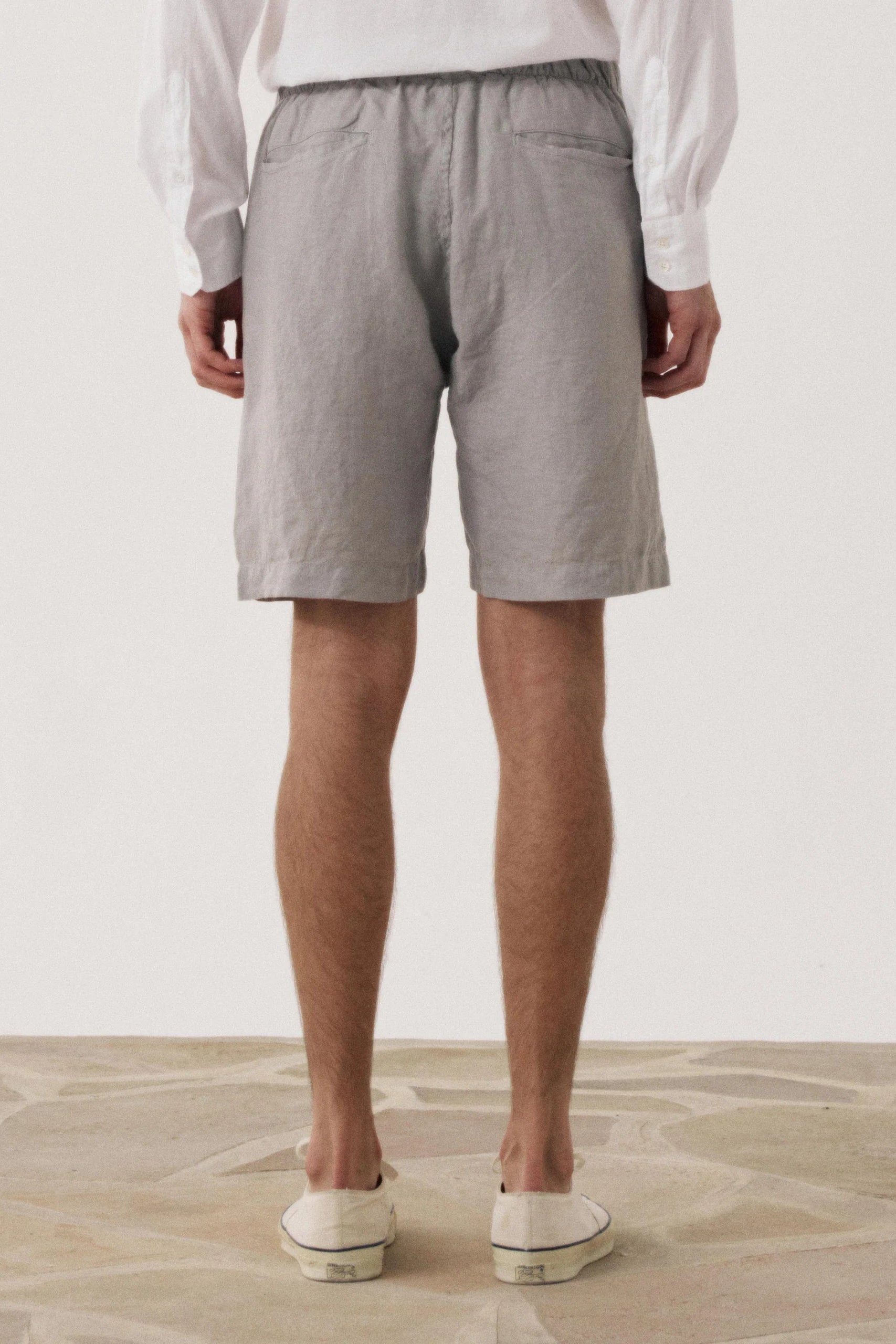 A person shown from the back wearing light gray linen shorts, a white long-sleeve shirt, and white slip-on shoes, standing against a plain off-white wall.