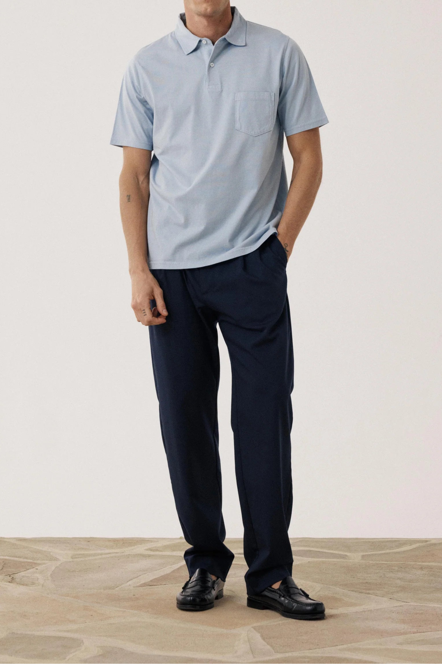 Man wearing a light blue polo shirt and dark pants standing against a plain background