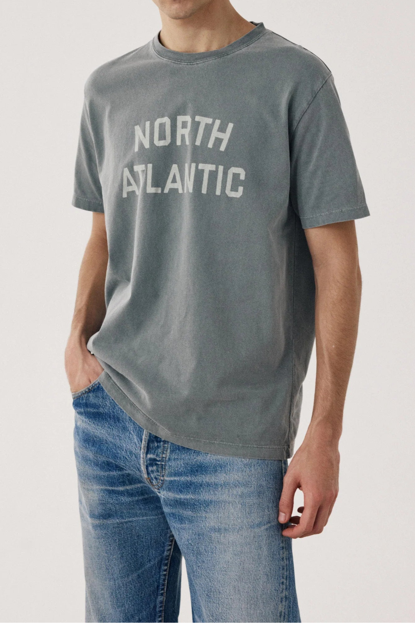 A man wearing a gray short-sleeve t-shirt with 'NORTH ATLANTIC' printed in white across the chest, paired with blue jeans, against a plain light gray studio background.