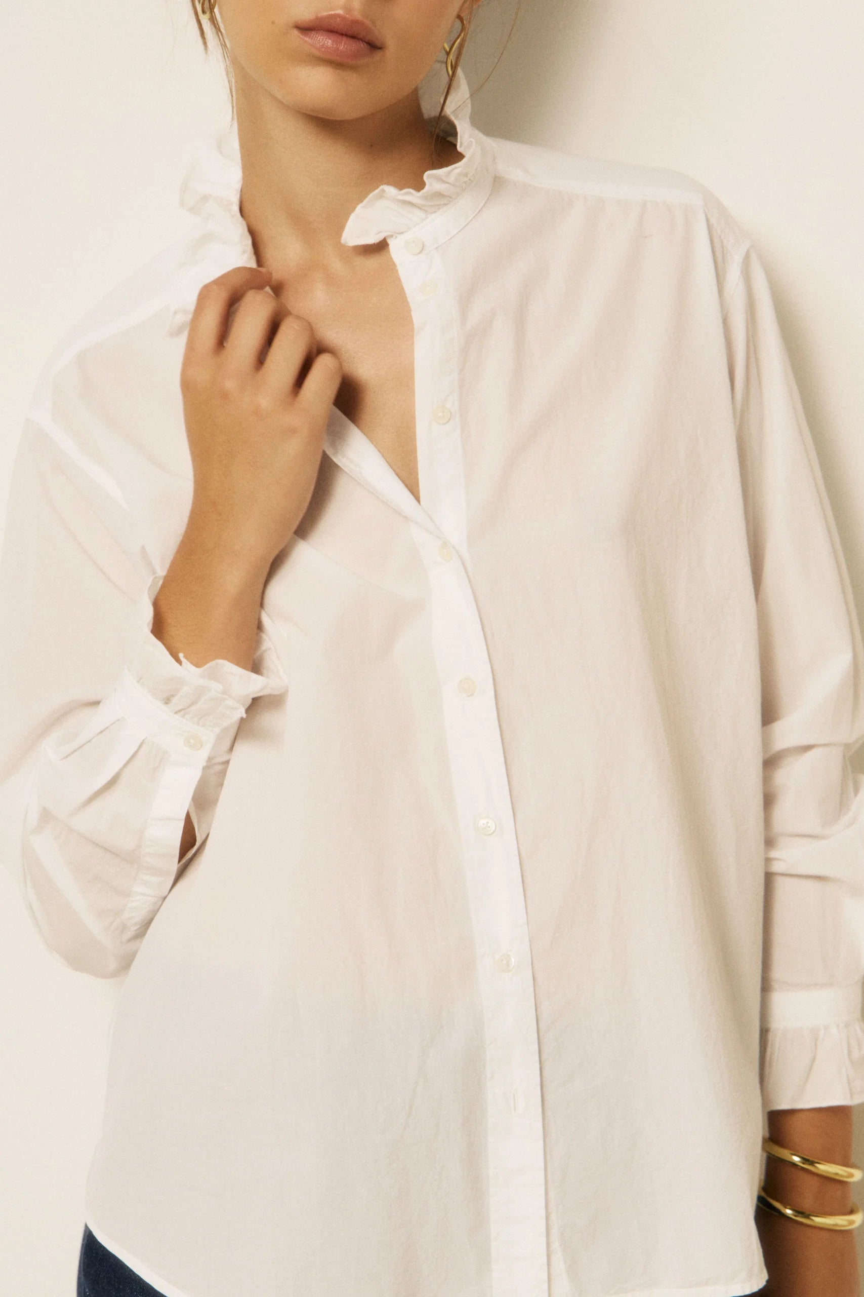 A woman wearing a white long-sleeve button-up shirt with a ruffled collar and ruffled cuffs, shown from the neck down against a plain light background.