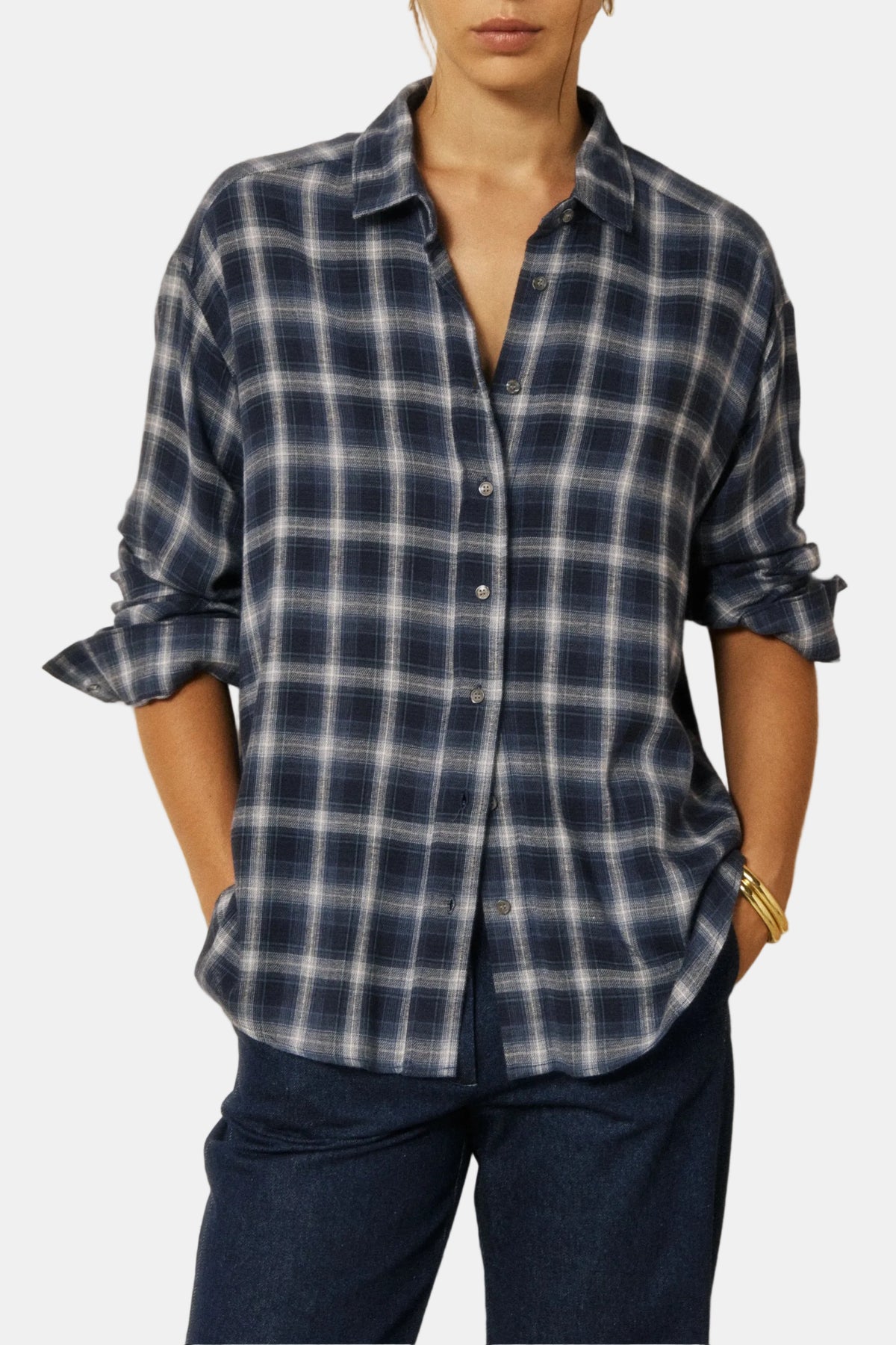 A woman wearing a navy and blue plaid button-down shirt with dark blue jeans, standing against a split cream and wood-paneled wall.