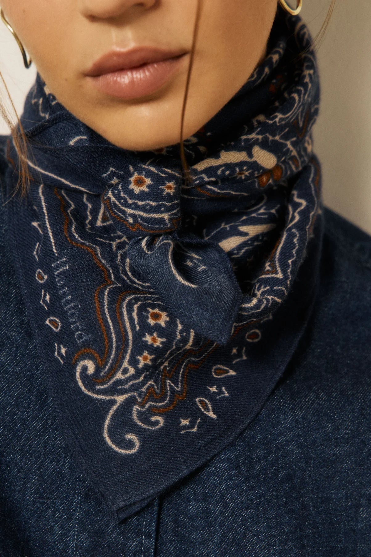 A close-up of a model wearing a navy blue paisley bandana scarf tied around their neck, paired with a dark blue denim shirt. The scarf features cream and brown paisley patterns with small embroidered details.