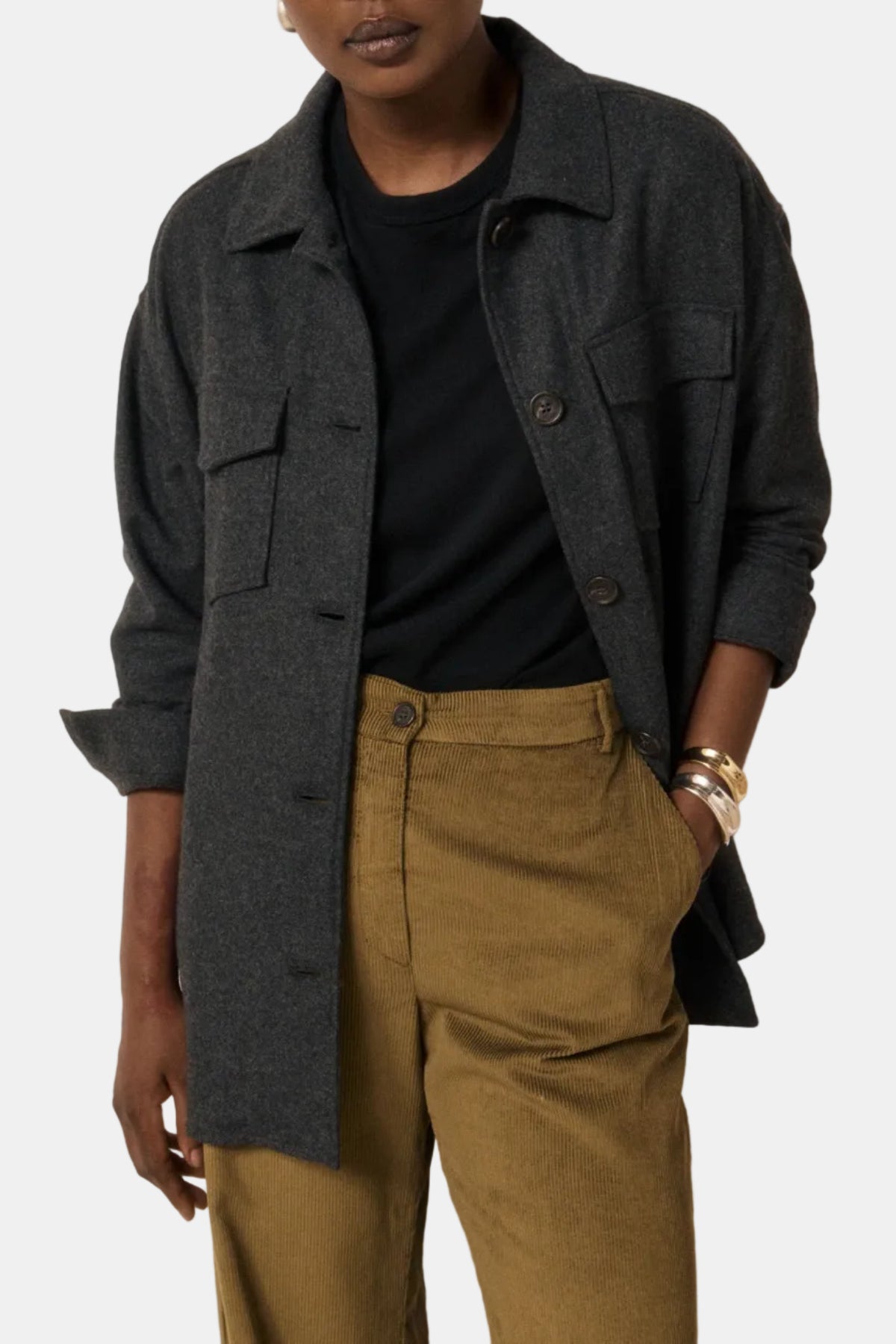 A model wearing a dark gray textured overshirt over a black t-shirt and olive green trousers, standing against a plain off-white wall.
