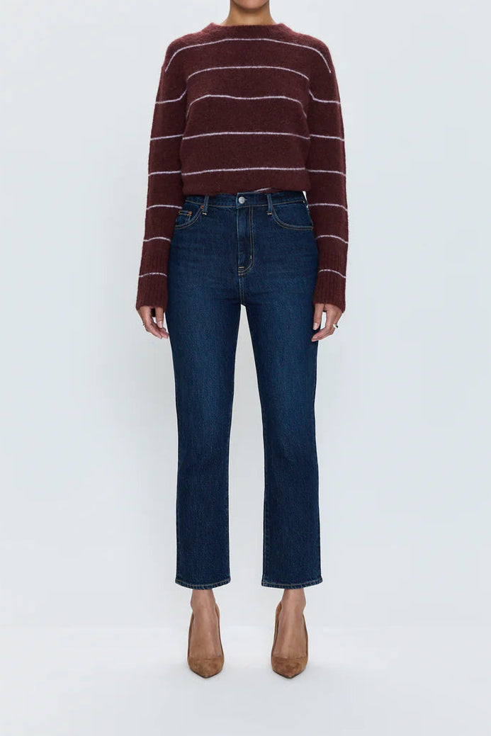 A model stands against a light gray studio background wearing a burgundy and white striped knit sweater, dark blue straight-leg jeans, and tan pointed-toe heels.