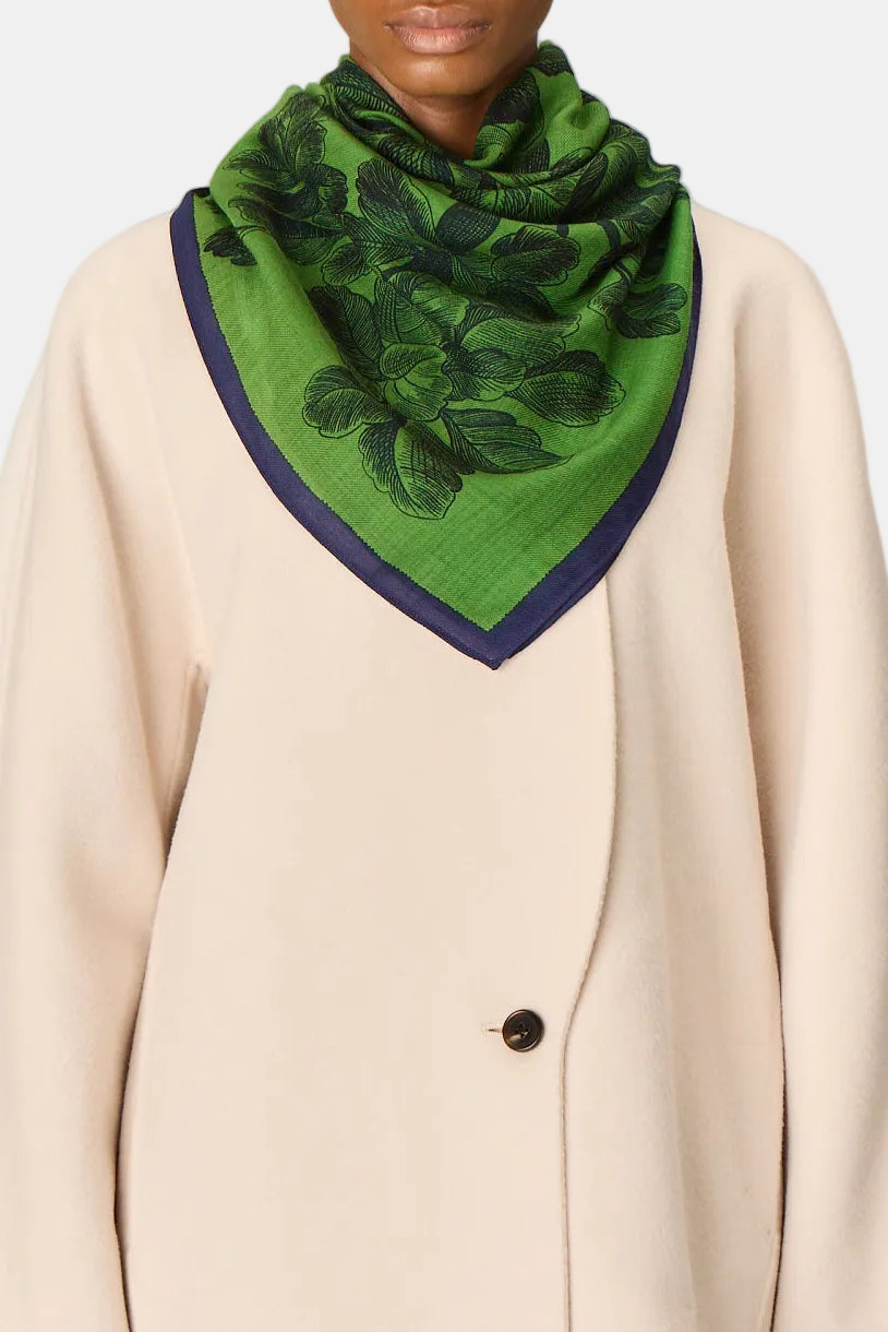 A model wearing a cream-colored double-breasted blazer and matching trousers, styled with a green patterned silk scarf featuring a dark green leaf design and a navy blue border, set against a plain light gray studio background.