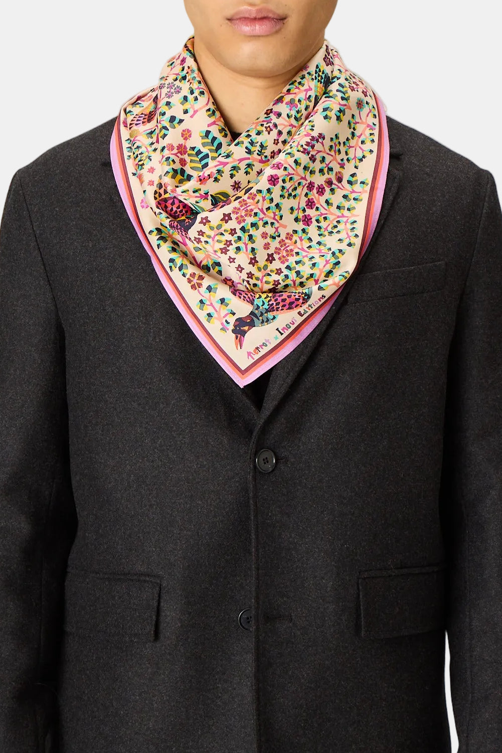 A man wearing a dark charcoal gray blazer and a colorful patterned neck scarf with pink trim, standing against a solid light gray background.