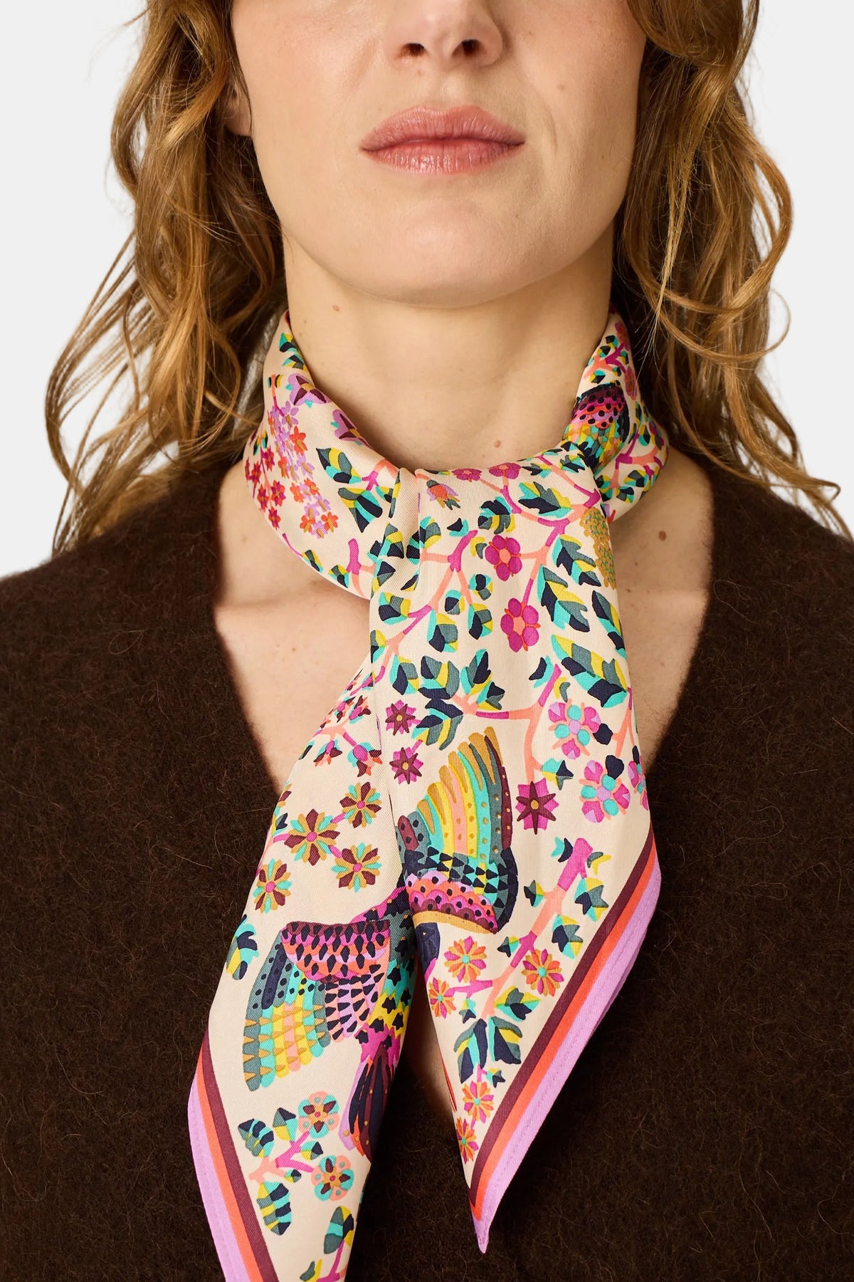 A close-up of a model wearing a cream-colored silk scarf with a vibrant multicolor floral and bird pattern, styled around the neck over a brown knit sweater.
