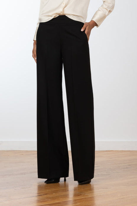 A woman stands in a studio wearing a cream satin button-up blouse and black wide-leg trousers, photographed against a light gray wall and wood floor.