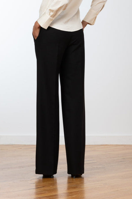 A woman wearing a cream long-sleeve blouse and black wide-leg trousers, shown from the back in a studio setting with a white wall and light wood floor.