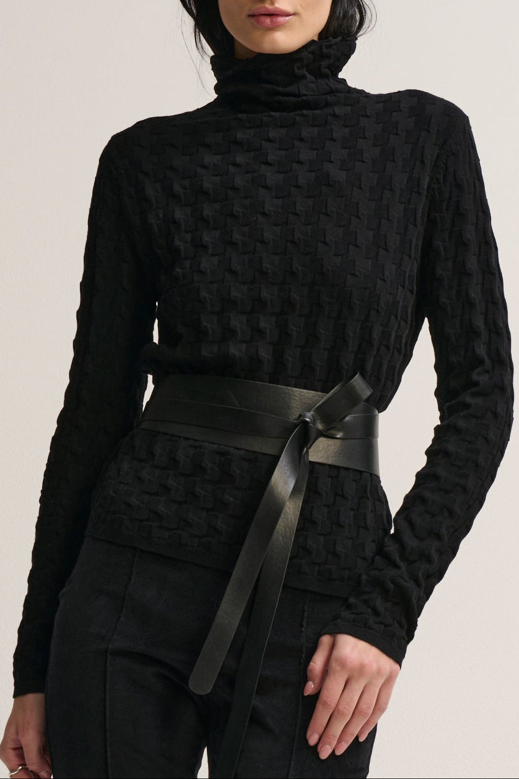 Woman wearing a black textured sweater with a belt against a neutral background