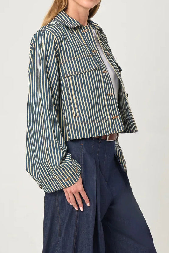A woman modeling a cropped, long-sleeve striped button-up shirt in green and white, paired with dark wide-leg denim pants, against a plain light gray studio background.