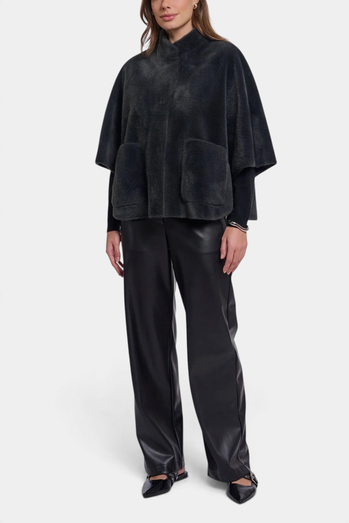 A model stands against a light gray studio background wearing a black textured cape-style jacket with a high collar and front patch pockets, paired with black wide-leg pants and black pointed flats.
