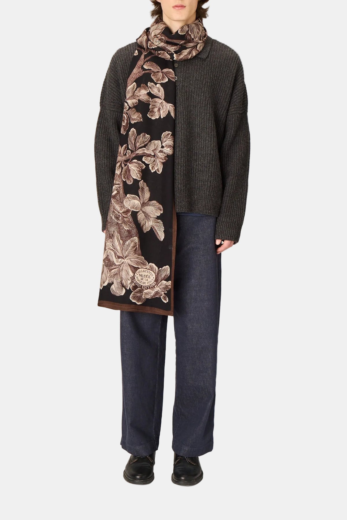 Person wearing a dark floral scarf, gray sweater, and blue pants on a plain background