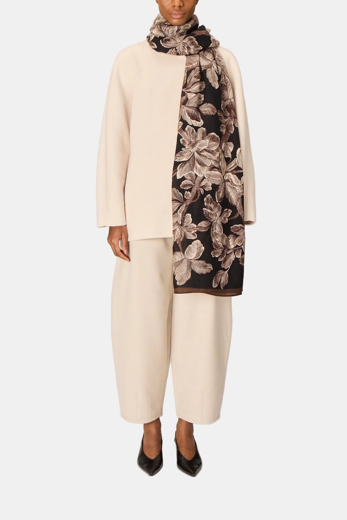 Woman wearing a light pink coat, white pants, and a floral scarf on a plain background