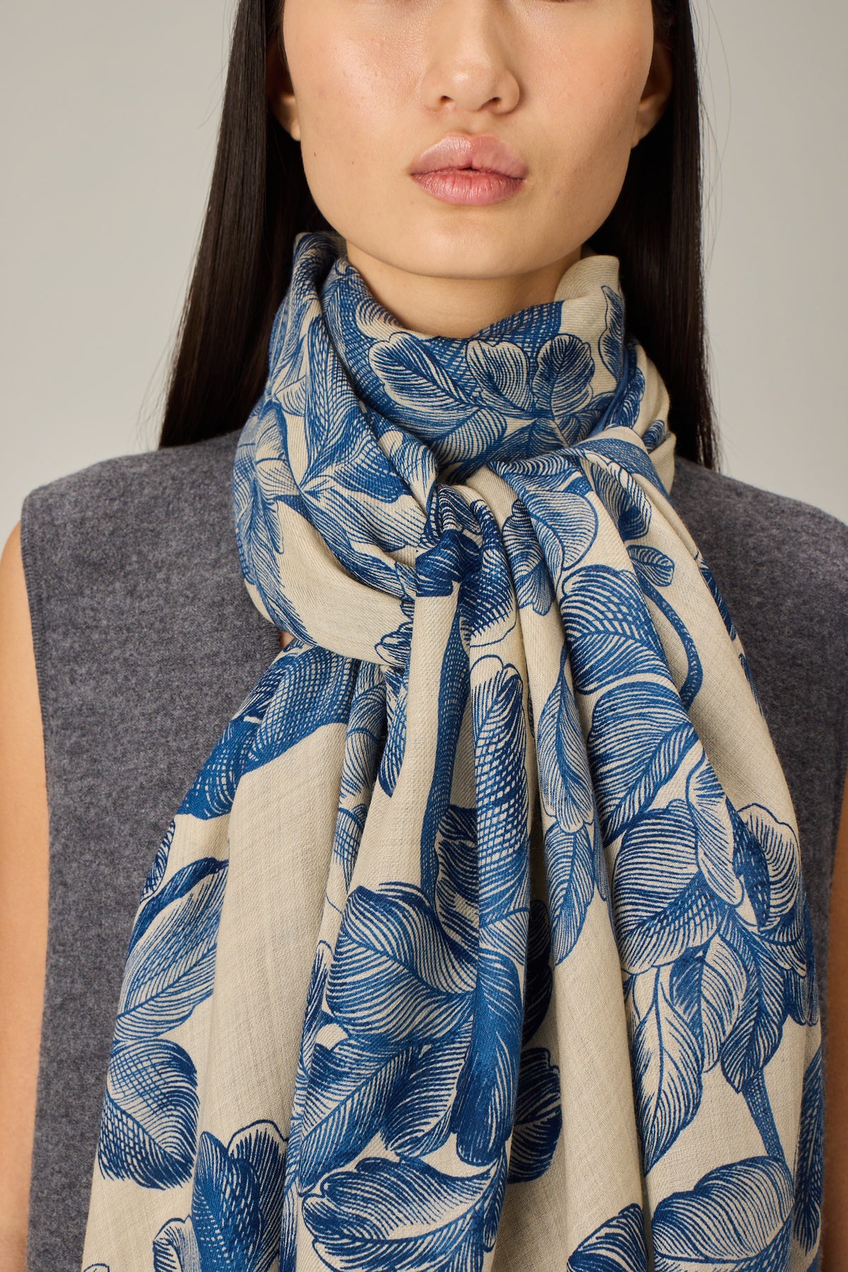 A close-up of a model wearing a light beige scarf with a blue botanical leaf pattern, styled over a gray sleeveless top against a neutral gray background.